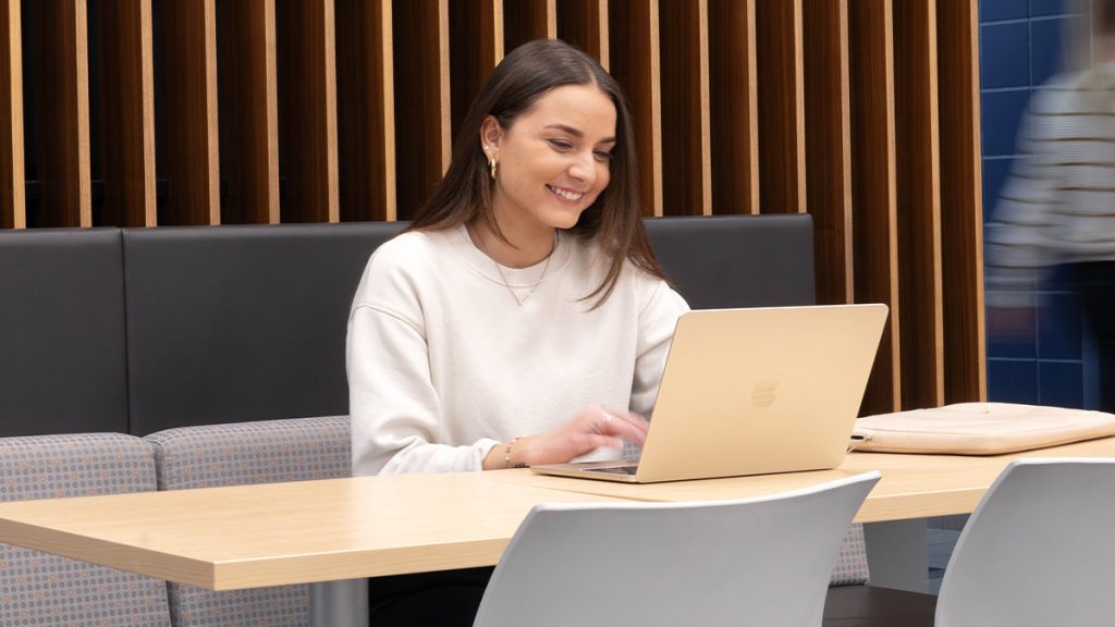 Une étudiante souriante rédige un texte sur son ordinateur portable dans un espace d’étude à l’UQAM.